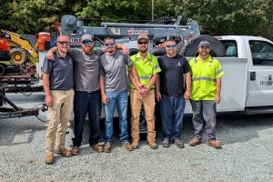 Six town employees stand shoulder to shoulder in front of public works equipment and trucks