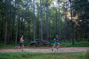 Two people run past an old racecar while running the grit trail with trees in the background