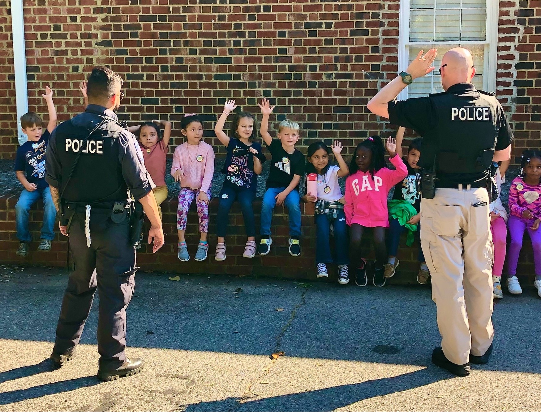 Two officers talking with children sitting behind brick police building